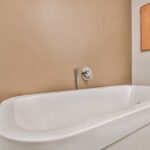 Big white soaking tub with wall mount chrome faucet and shower nozzle at beige wall in bathroom with painting on wall