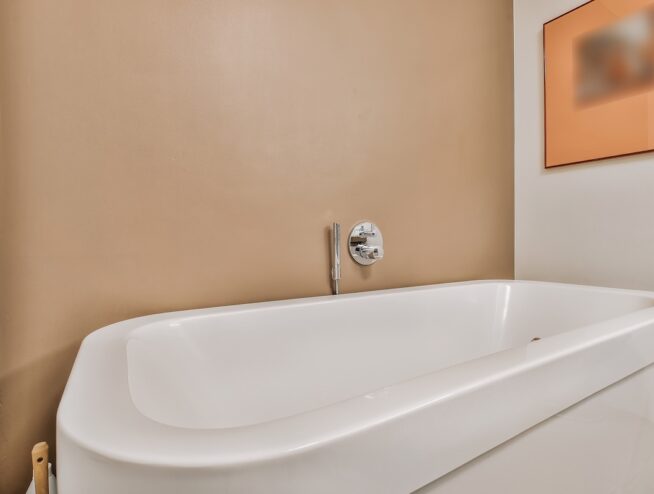 Big white soaking tub with wall mount chrome faucet and shower nozzle at beige wall in bathroom with painting on wall