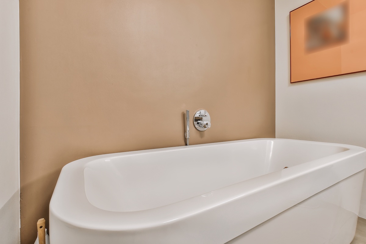 Big white soaking tub with wall mount chrome faucet and shower nozzle at beige wall in bathroom with painting on wall